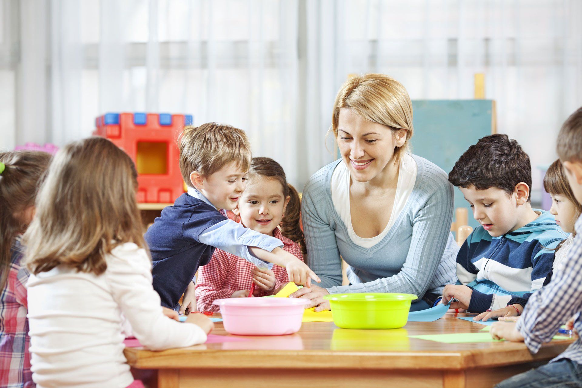 A teacher and group of young children sitting at a table with art supplies, many with their hands raised.