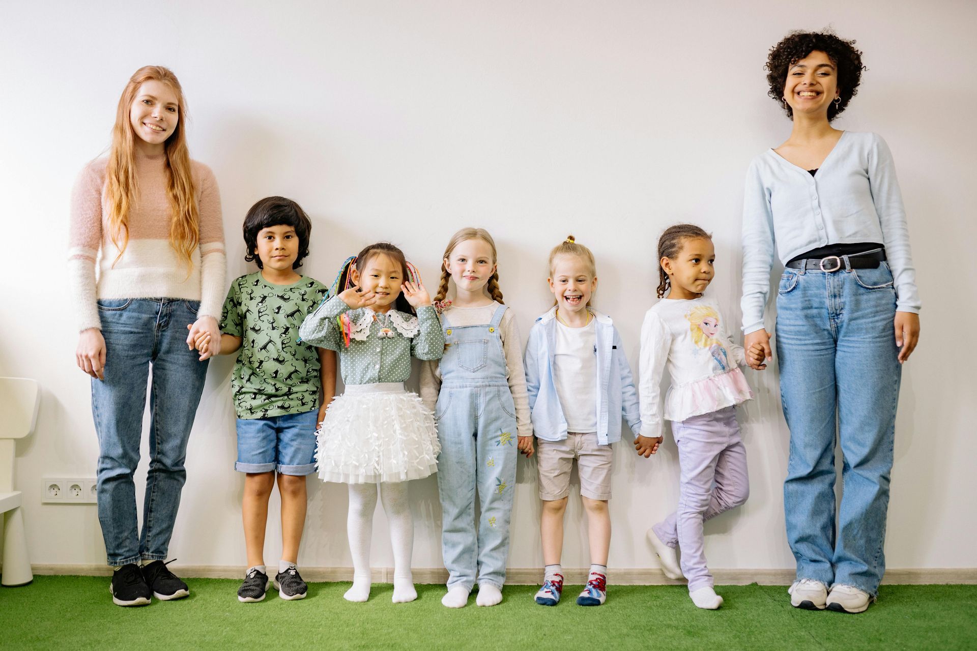 Two adults and five children stand in a line against a white wall over a green floor, smiling for a group photo.