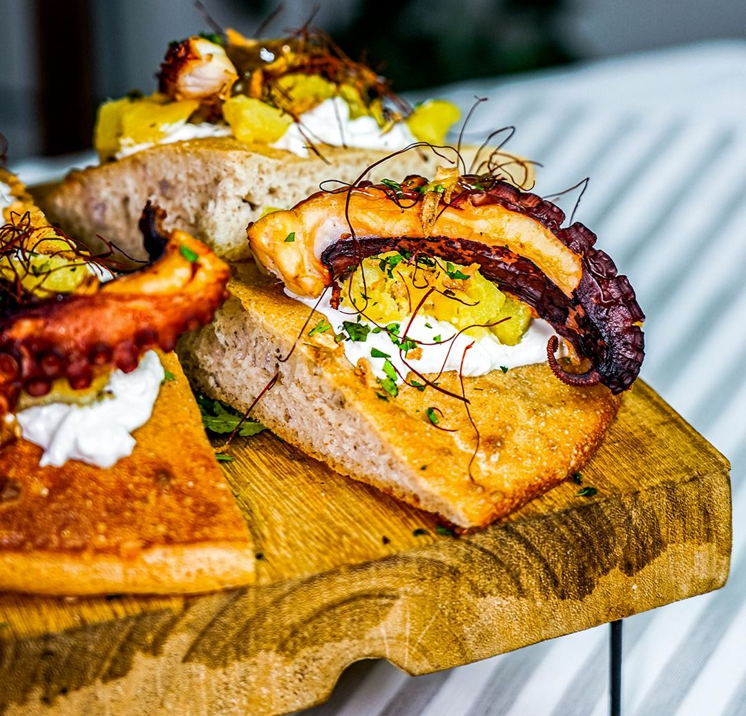 Polpo grigliato su pane tostato, con panna e guarnizione, servito su un tagliere di legno.