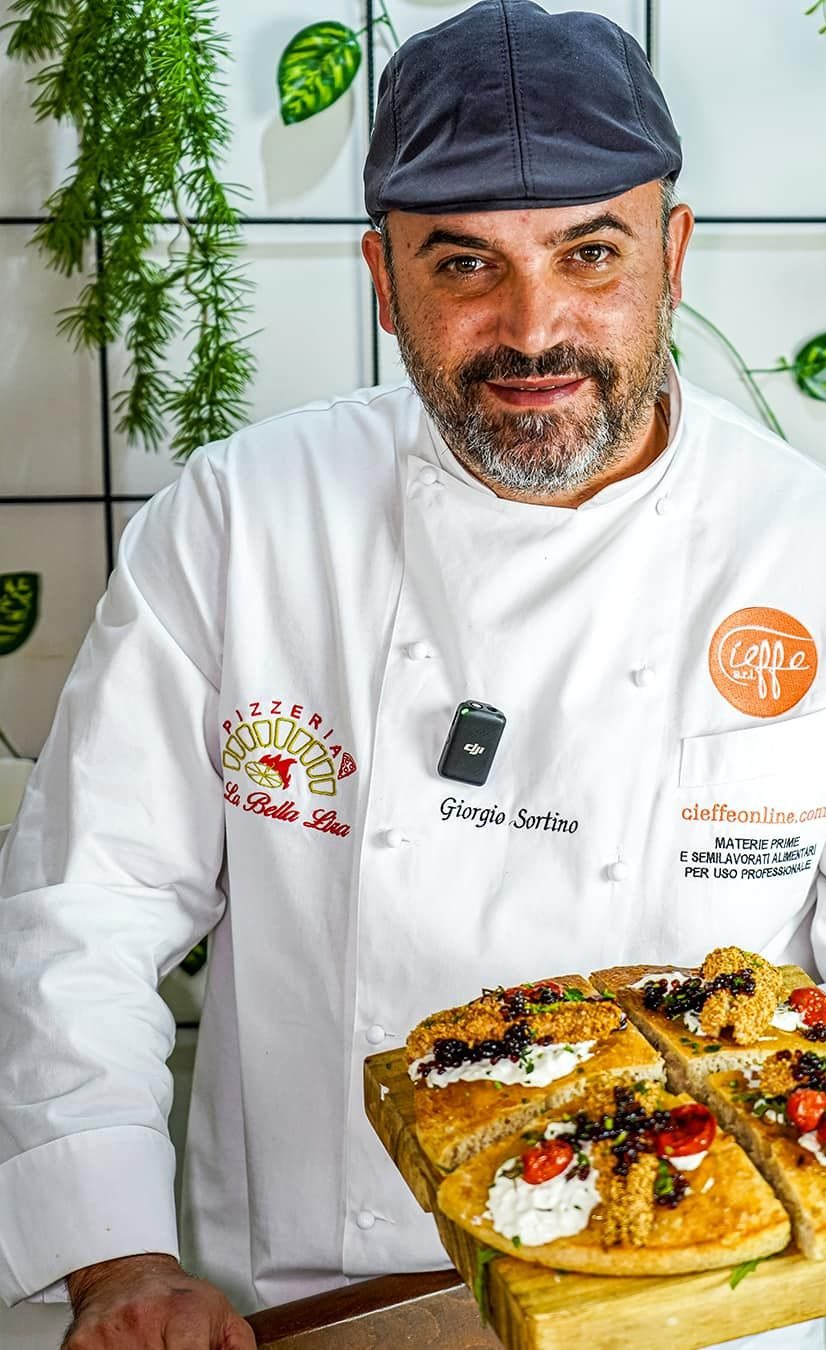 Cuoco che tiene in mano una focaccia condita, sorridente, con indosso un cappello e un cappotto da cuoco, all'interno di una cucina.