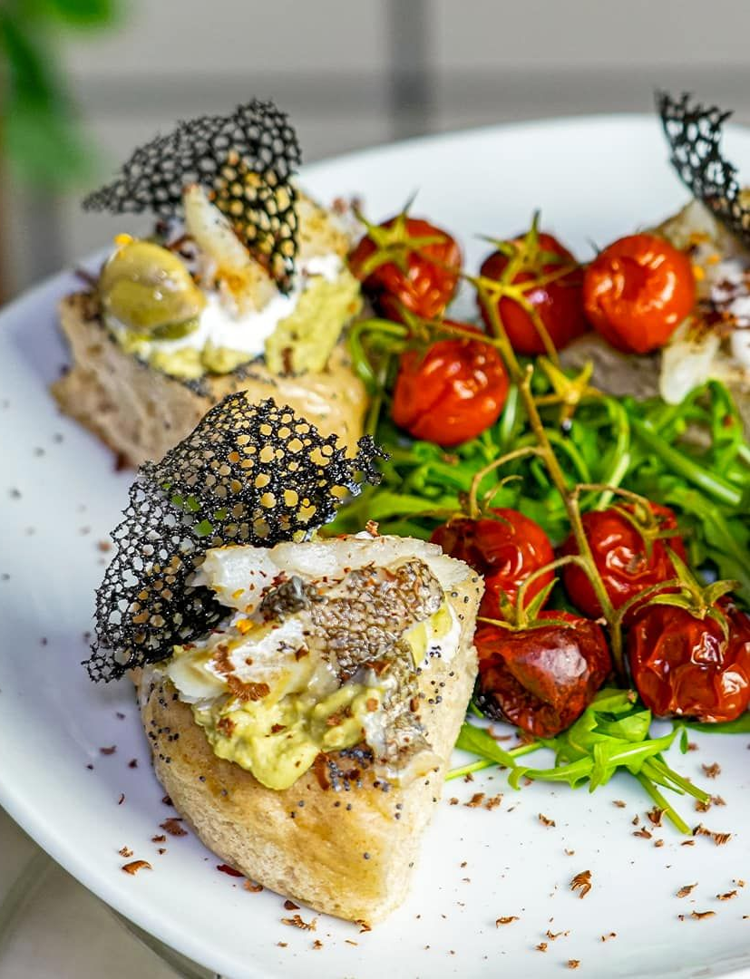 Pane tostato con avocado, formaggio e pomodori, servito con rucola e guarnizione nera.