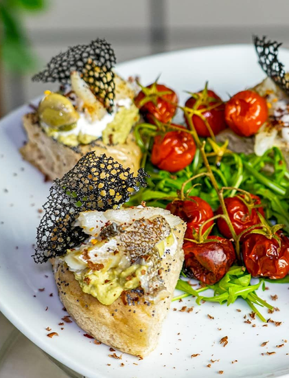 Pane tostato con avocado, formaggio e pomodori, servito con rucola e guarnizione nera.