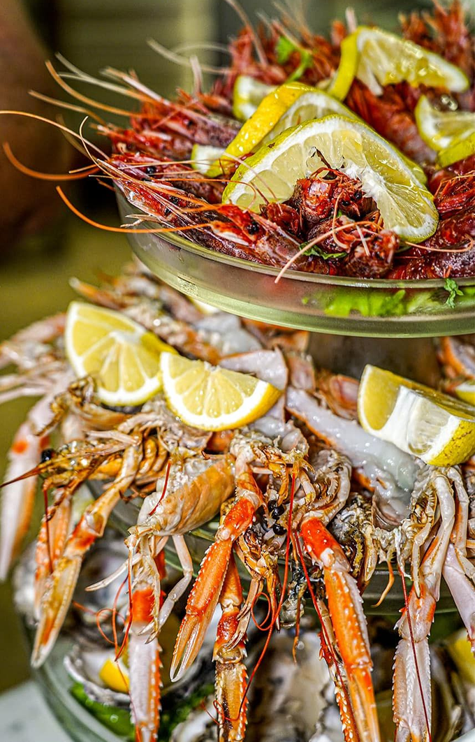 Torre di frutti di mare: vari gamberi e scampi cotti, disposti su vassoi di vetro a più livelli, guarnito con fette di limone.