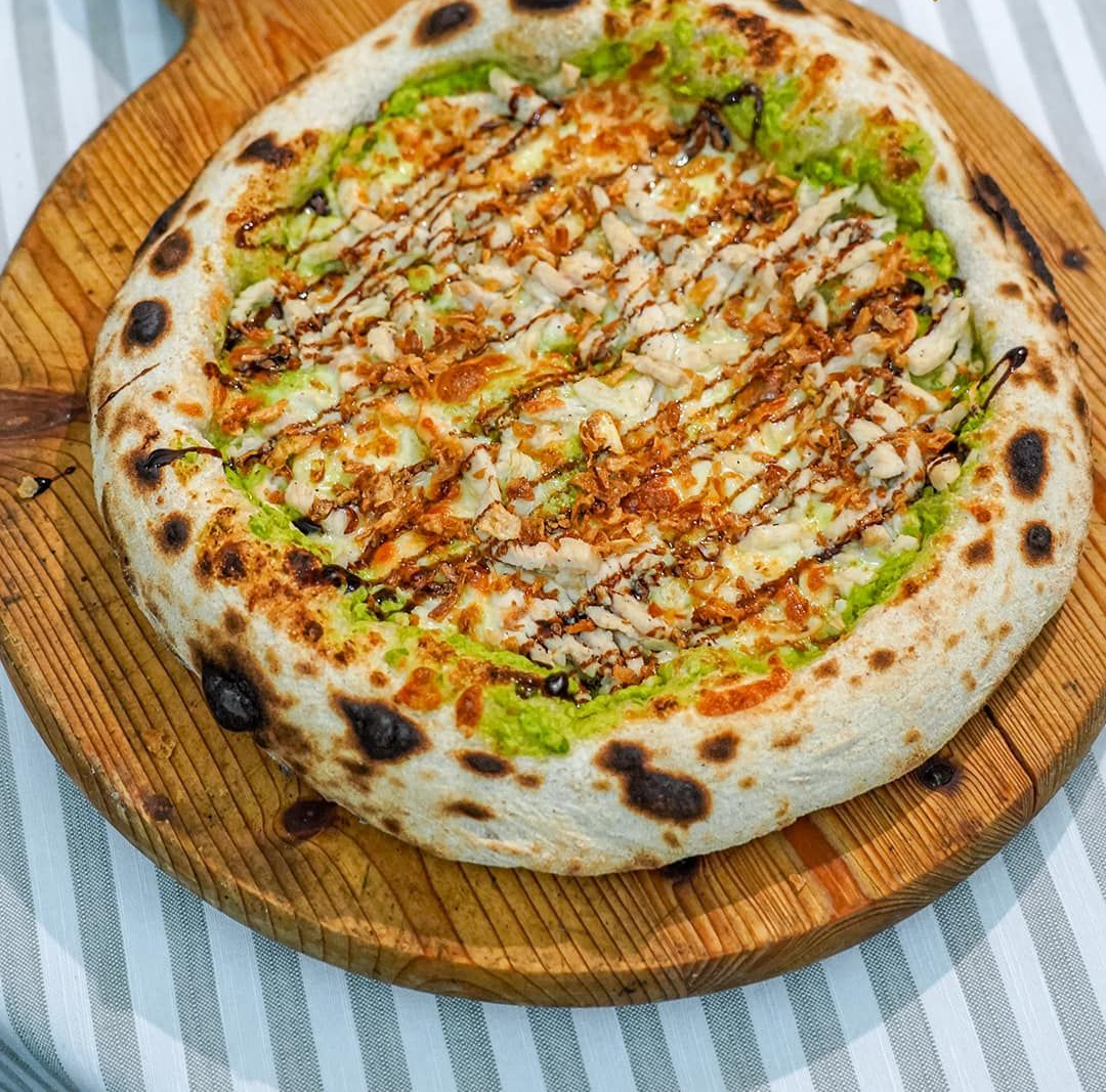 Pizza su tavola di legno; base verde, condimenti e crosta marrone. Salsa a filo.