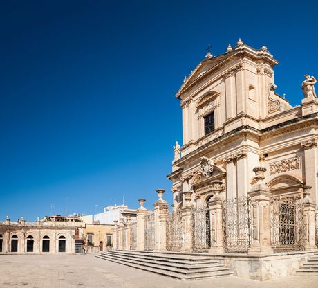 Facciata di una chiesa barocca contro un cielo azzurro terso; scale e un cancello decorativo di fronte.