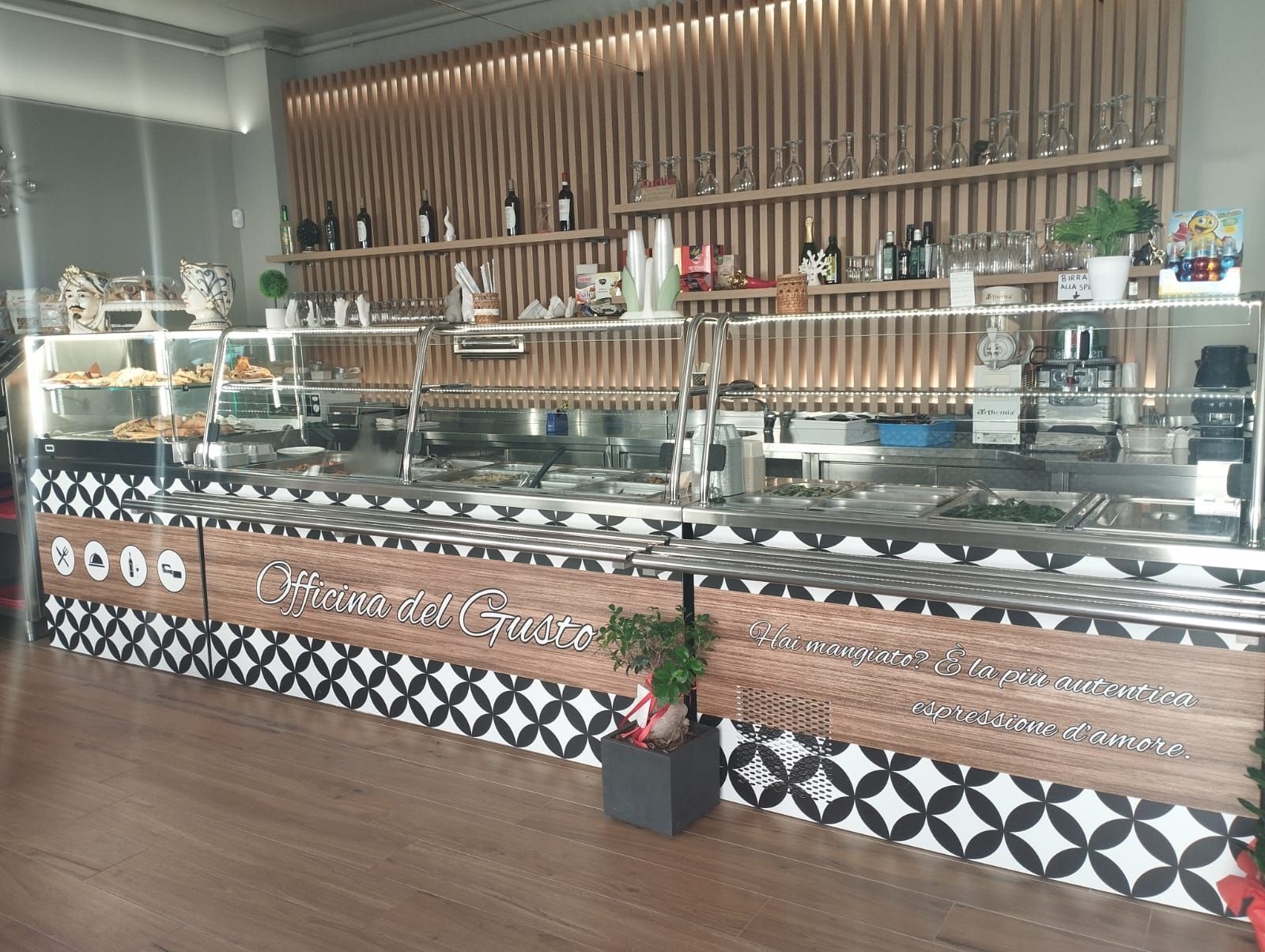 Bancone con esposizione di prodotti alimentari in un negozio, piastrelle marroni e bianche. Sfondo in legno con ripiani, vari articoli a vista.