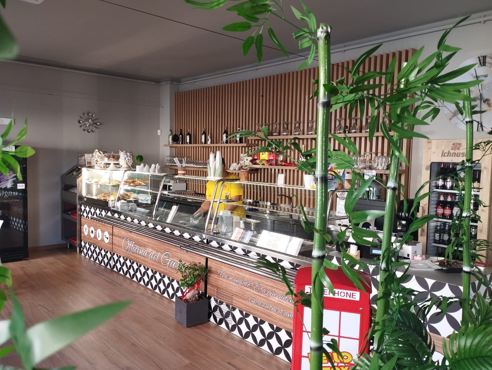 Interno di un bar con bancone con esposizione di cibo, parete con doghe in legno, piante di bambù e bevande in frigorifero.