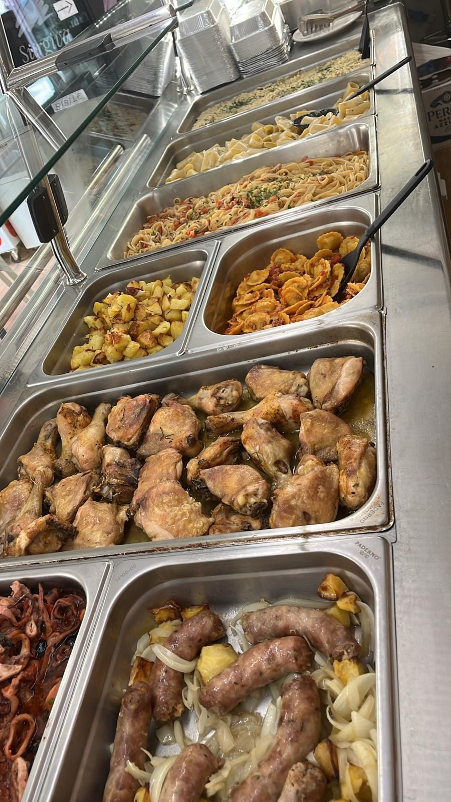 Buffet di cibo: pollo, patate, pasta e salsiccia esposti su vassoi di acciaio inossidabile sotto vetro.