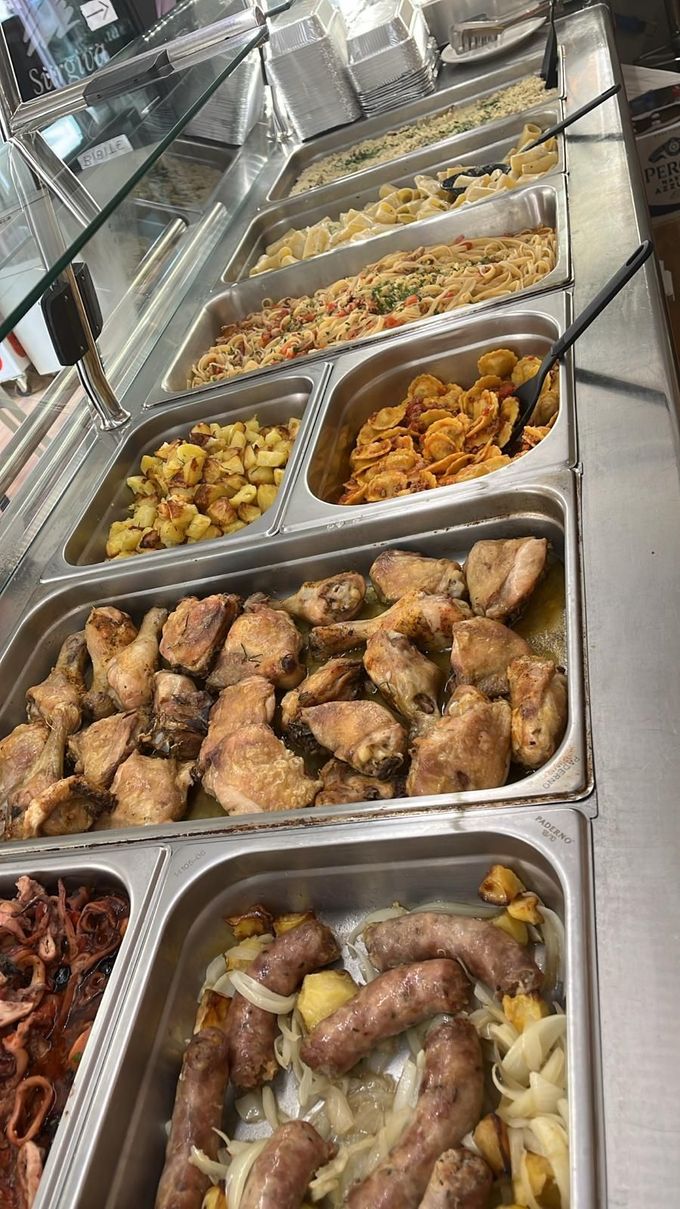 Buffet di cibo: pollo, patate, pasta e salsiccia esposti su vassoi di acciaio inossidabile sotto vetro.