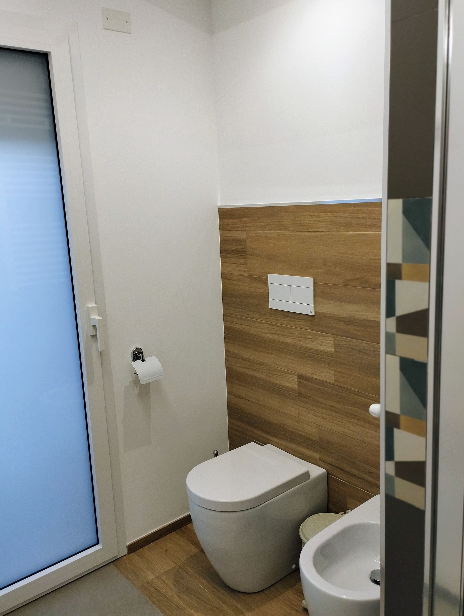 Bagno con WC bianco, bidet e parete rivestita in legno. Finestra smerigliata sulla sinistra.