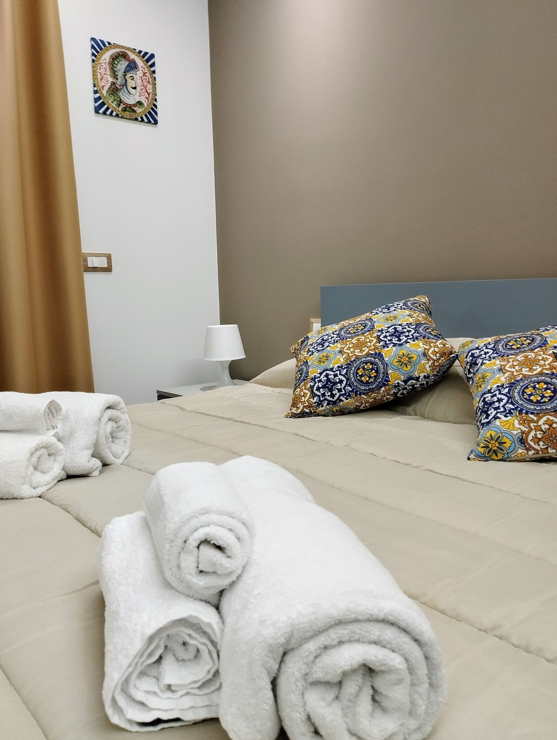 Camera da letto con asciugamani bianchi arrotolati sul letto, cuscini decorativi, un comodino e quadri alle pareti.