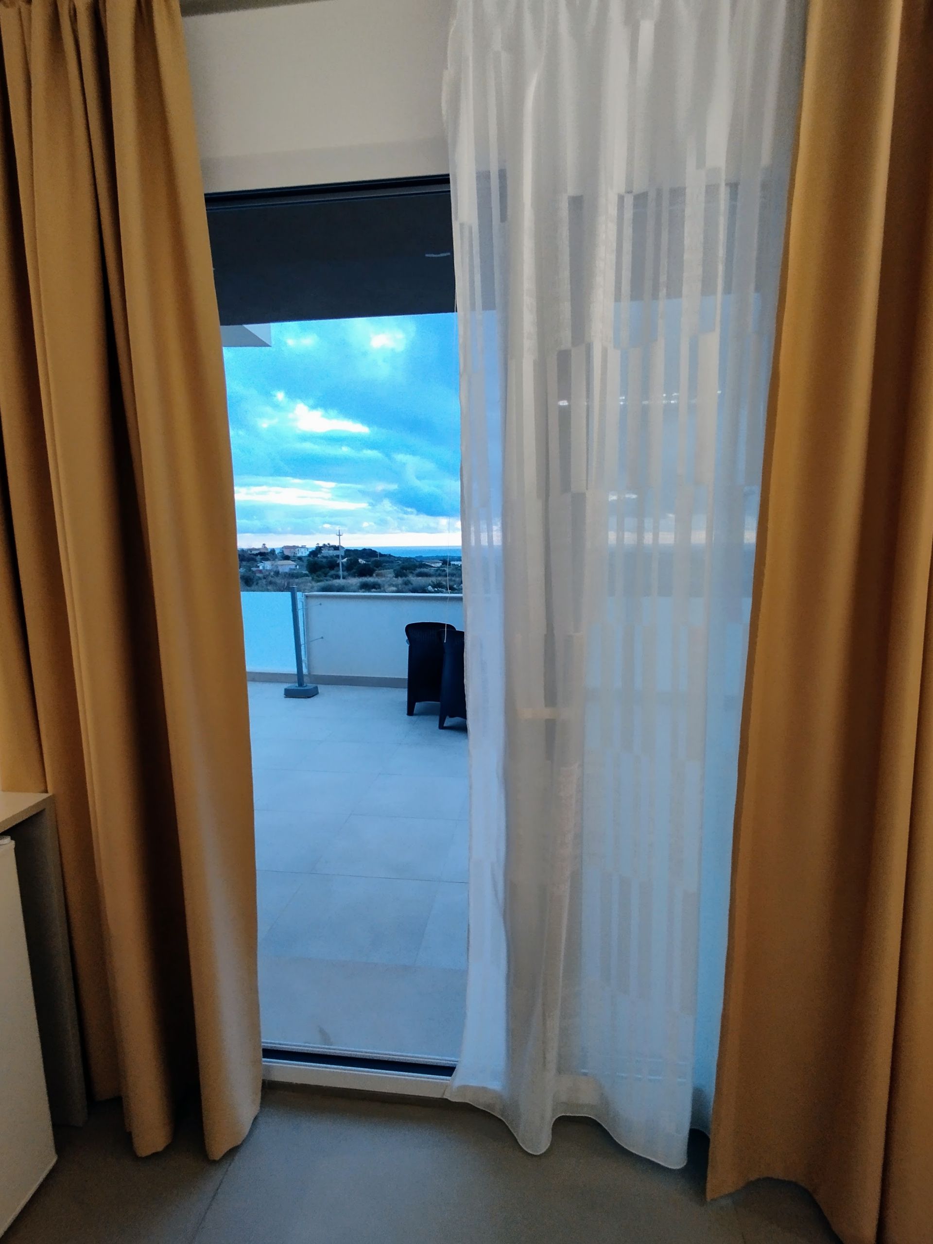 Vista di un balcone esterno attraverso una porta scorrevole in vetro aperta con tende bianche trasparenti e drappeggi dorati.