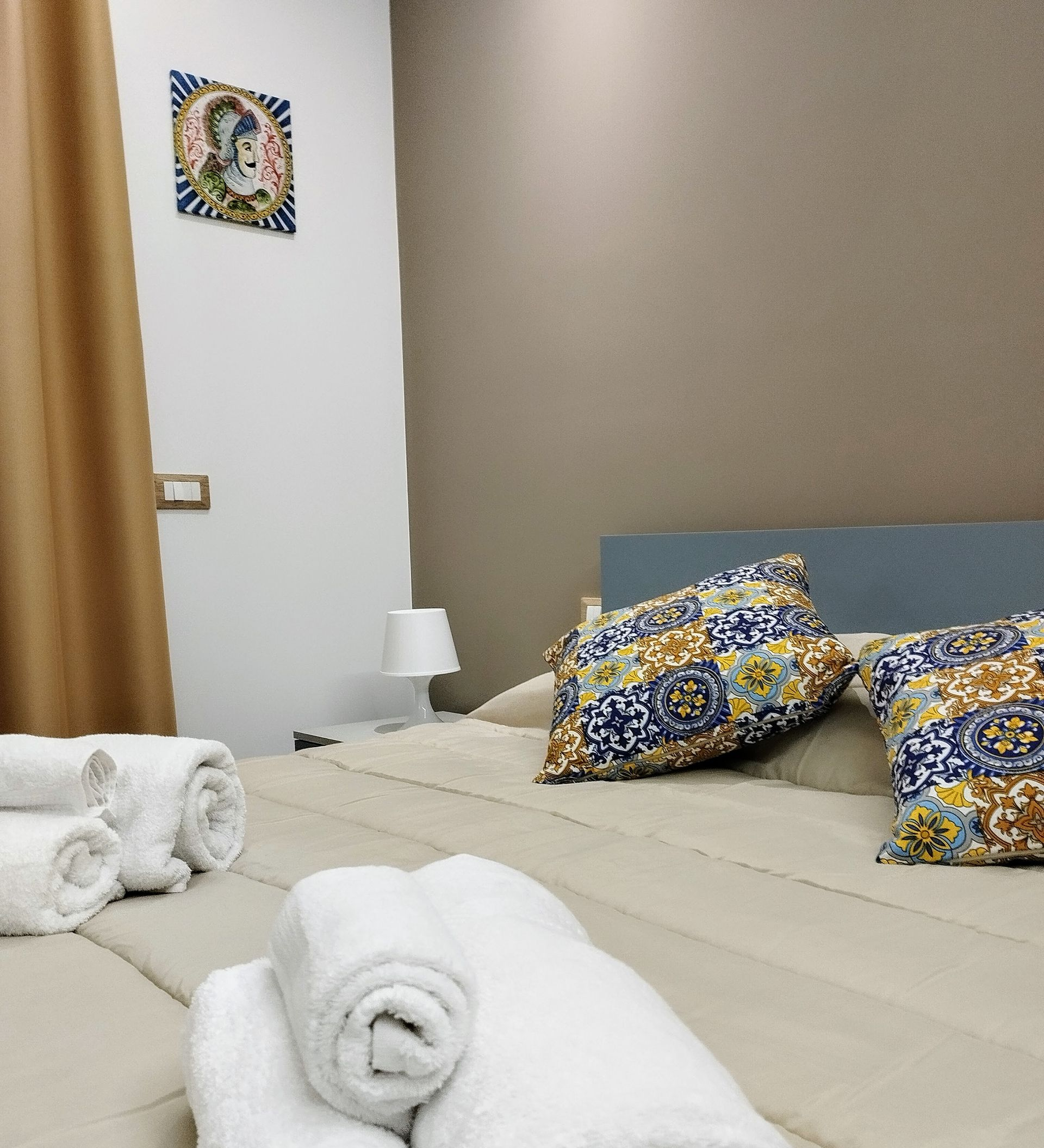 Letto con asciugamani bianchi arrotolati, cuscini, lampada e una piastrella decorativa sulla parete.