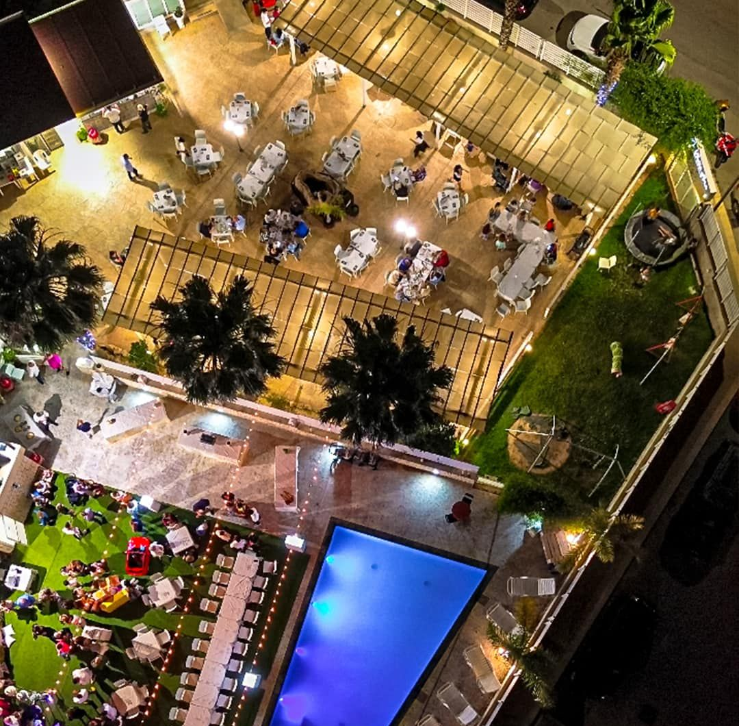 Vista dall'alto di un evento all'aperto illuminato con tavoli, piscina e spazio verde; sono presenti persone.