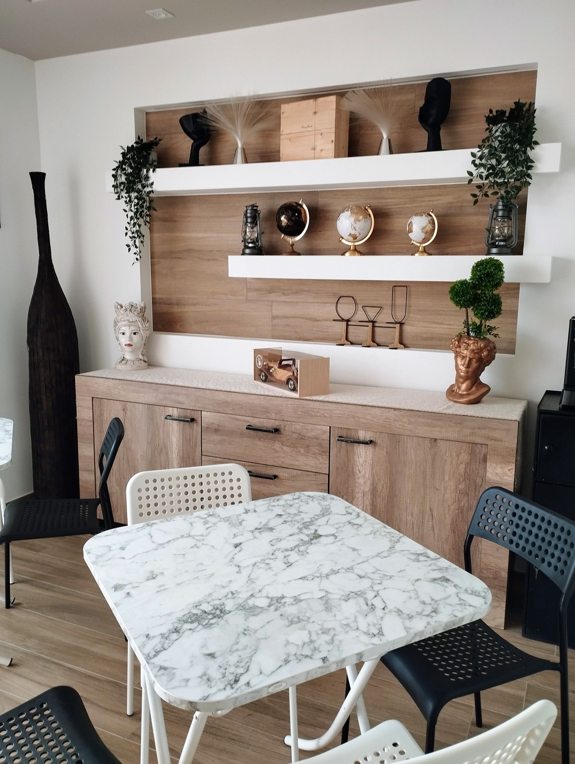 Zona pranzo con tavolo in marmo, credenza in legno, mensole con decorazioni, sedie bianche e nere.