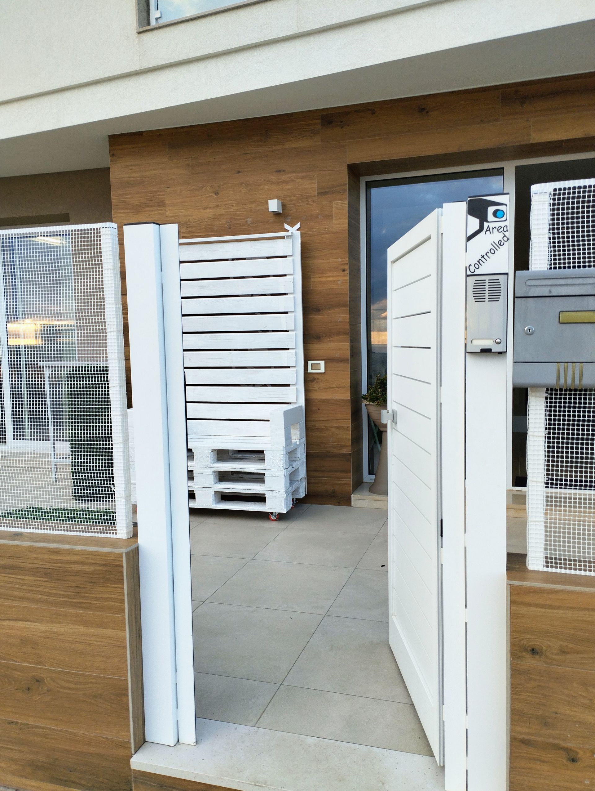 Cancello bianco che si apre su un portico con rivestimento effetto legno, una cassa di legno accatastata e una cassetta della posta.