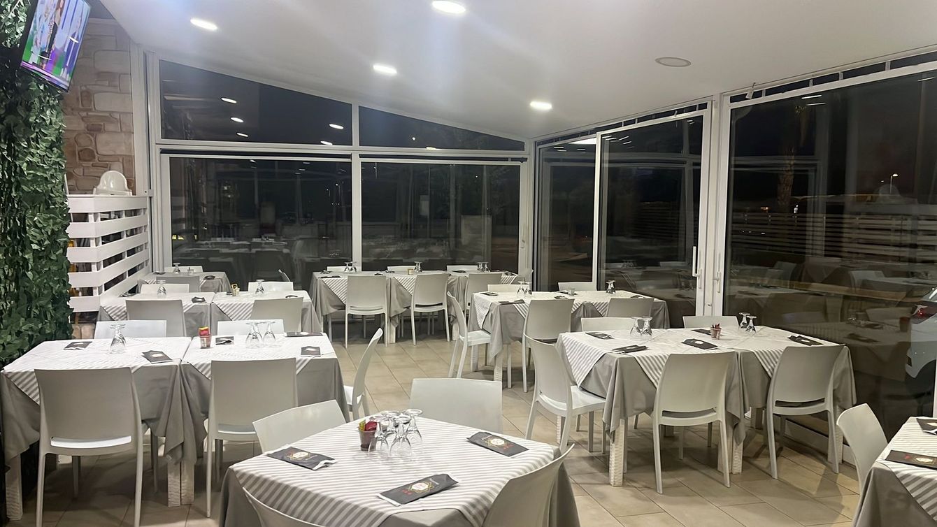 Interno di un ristorante con tavoli apparecchiati per cenare; sedie bianche, tovaglie neutre.