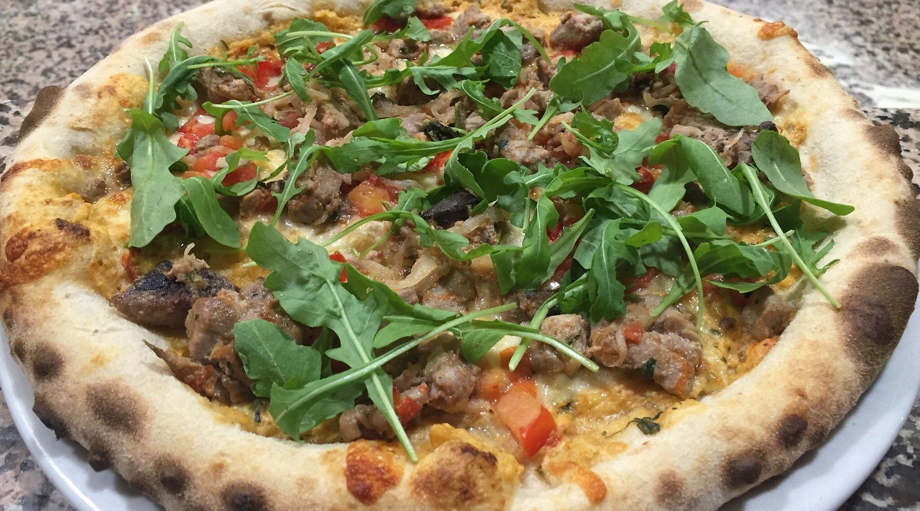 Pizza condita con rucola, salsiccia e pomodori su una base cotta nel forno a legna.