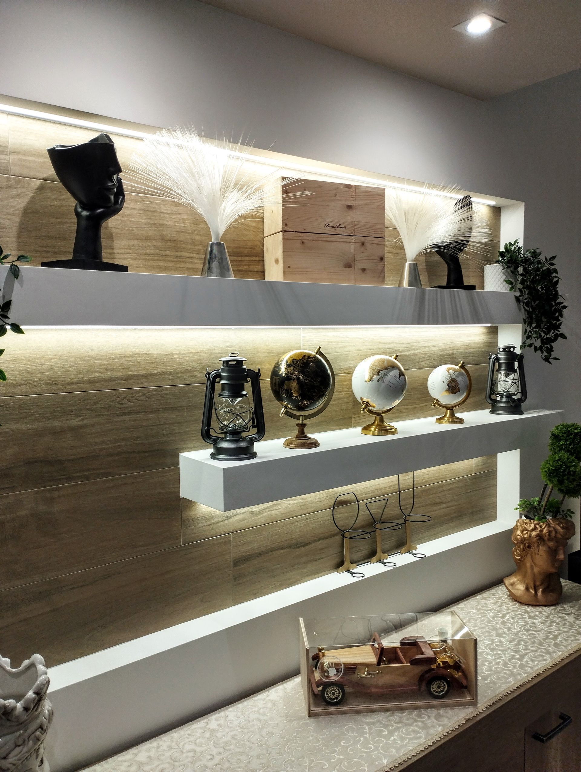 Mensole con pannello posteriore illuminato, che espongono globi decorativi, lampade e sculture in un interno moderno.
