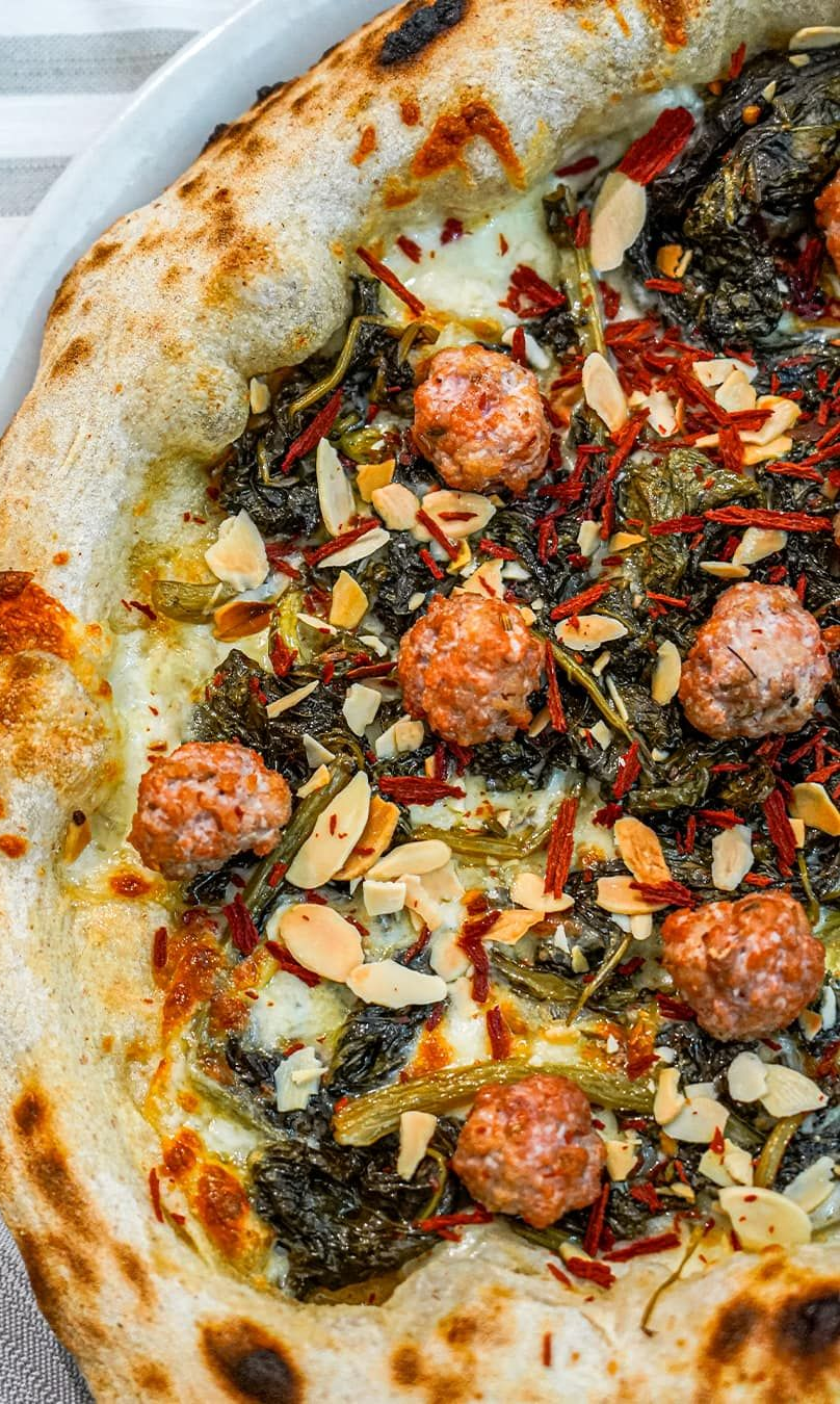 Pizza con salsiccia, verdure e mandorle su un piatto bianco.