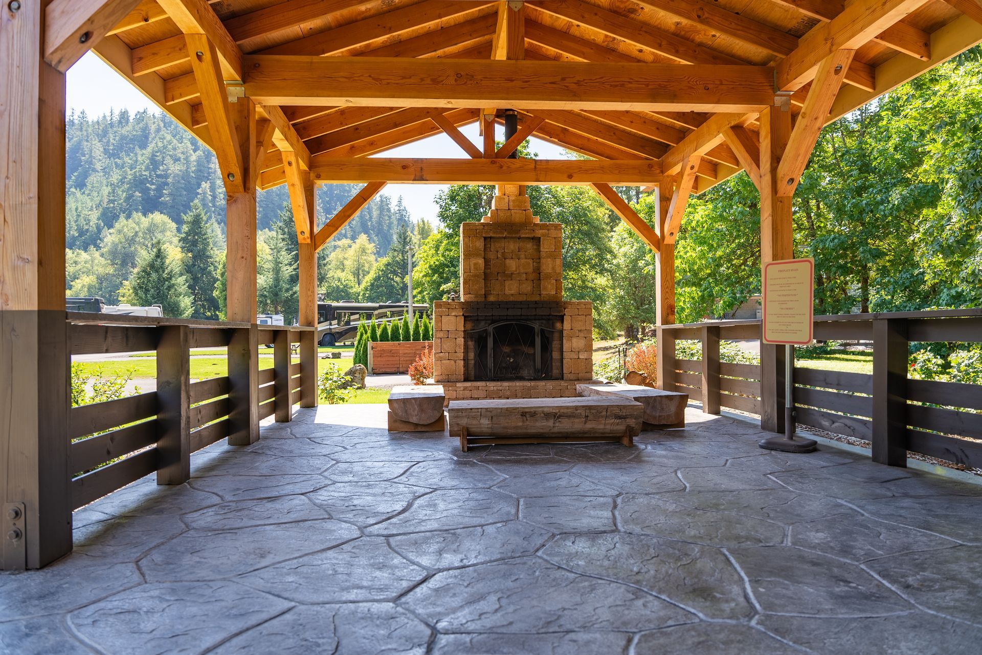 Wooden gazebo with stone fireplace, gray stamped concrete floor, and forest backdrop.