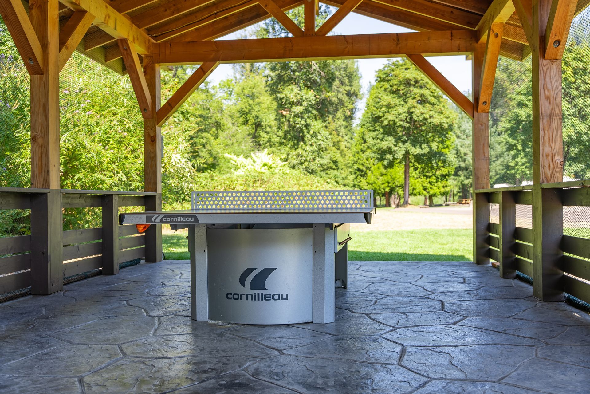 Ping pong table under a wooden gazebo in a park. Green trees and grass in the background.