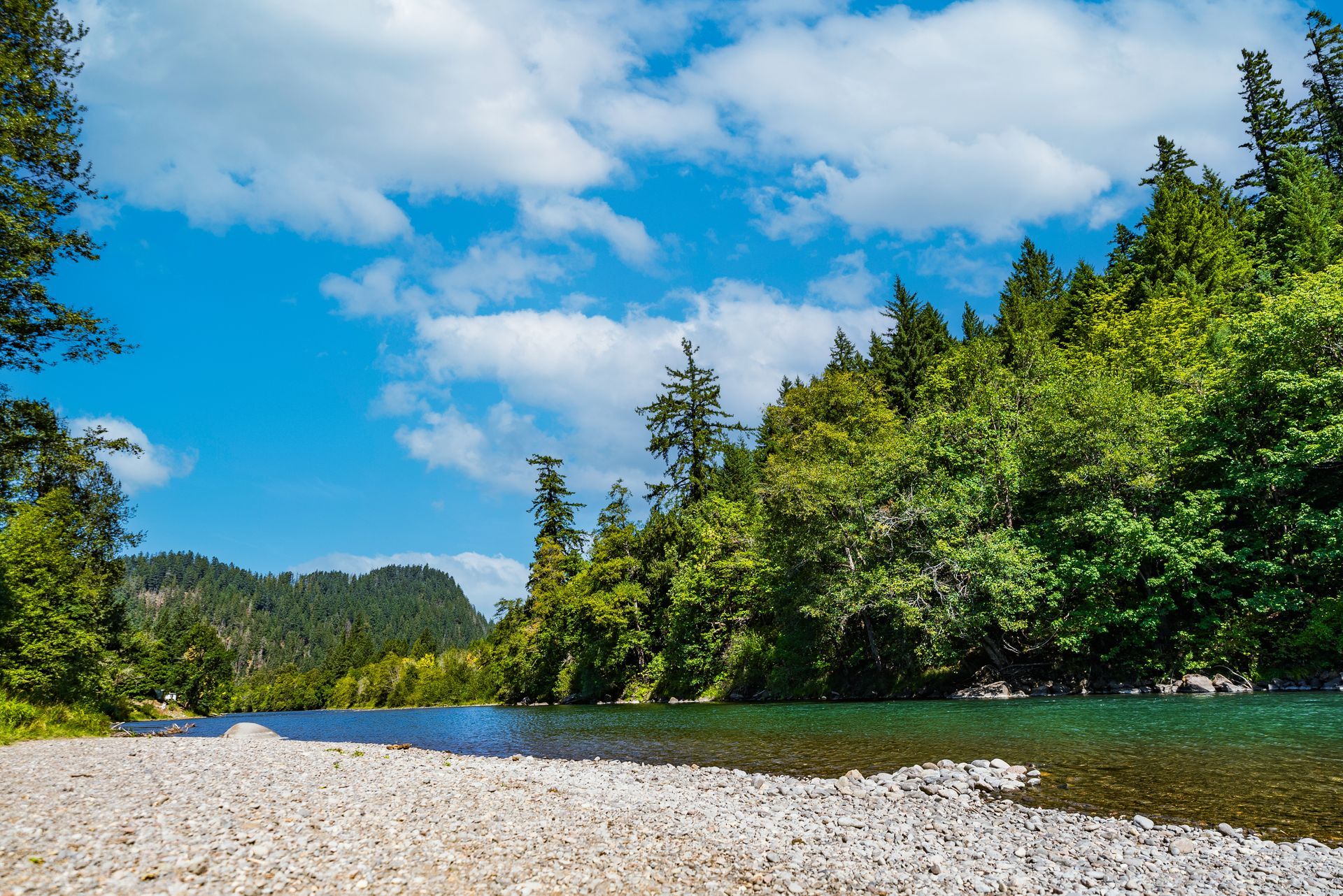Gravel shore with a turquoise river, lush green trees, and blue sky with clouds.