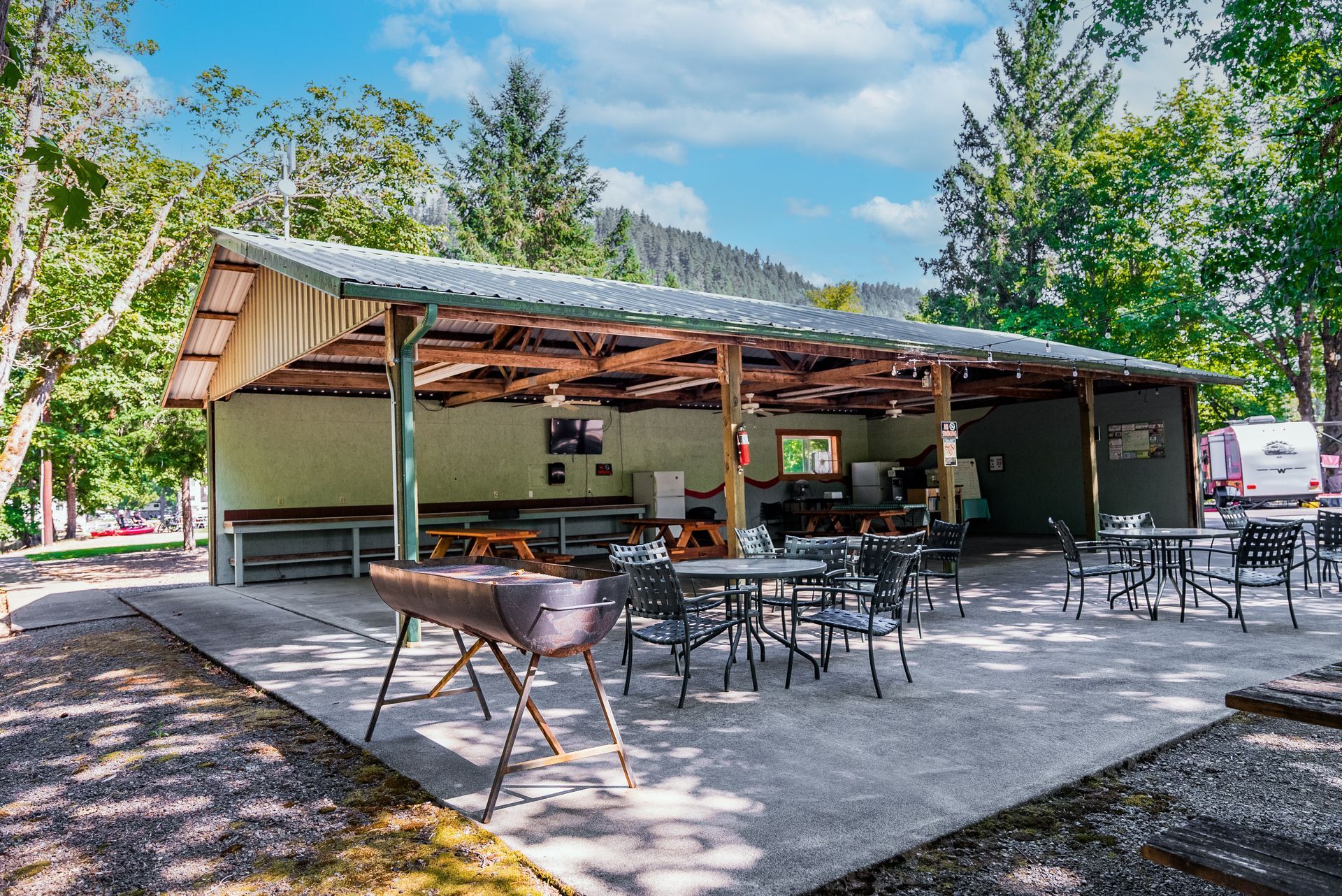 Open-air picnic shelter with tables, grill, and trees in background.