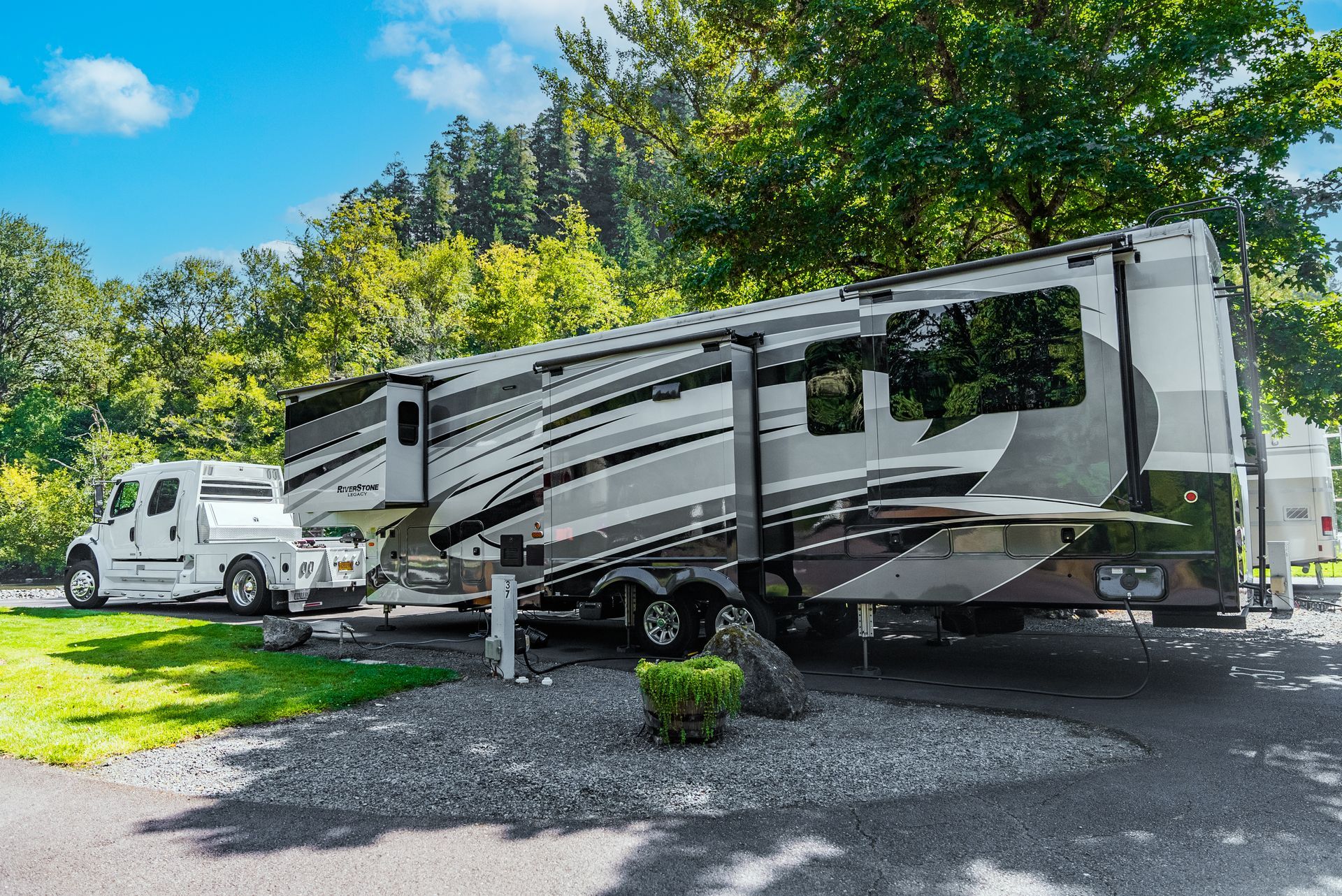 Large RV with a white truck parked in a gravel campsite, surrounded by grass and trees under a blue sky.