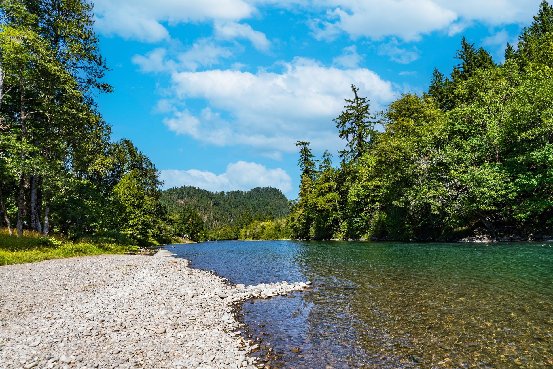 River shoreline with pebbles, calm water, and trees under a blue, cloudy sky.