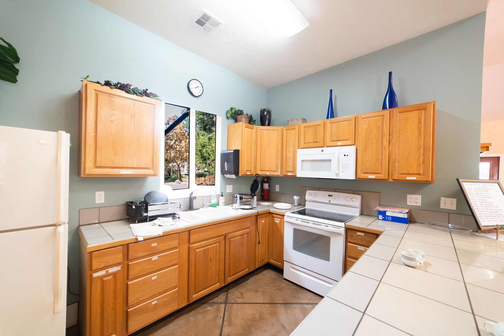 Kitchen with wooden cabinets, white appliances, window overlooking trees, and tile countertops.
