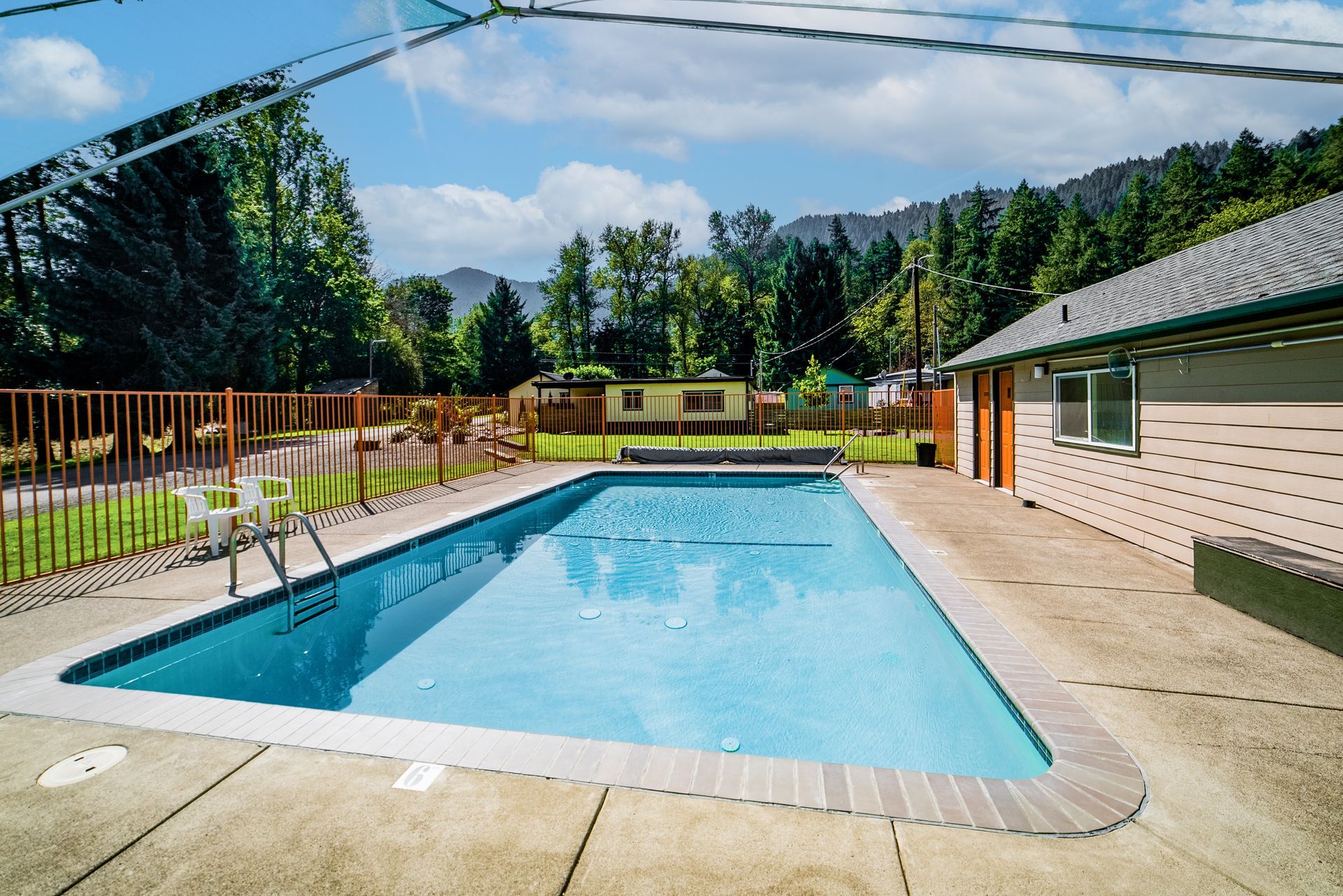 A rectangular swimming pool surrounded by a concrete patio and a low brown fence. The background has trees and mountains.