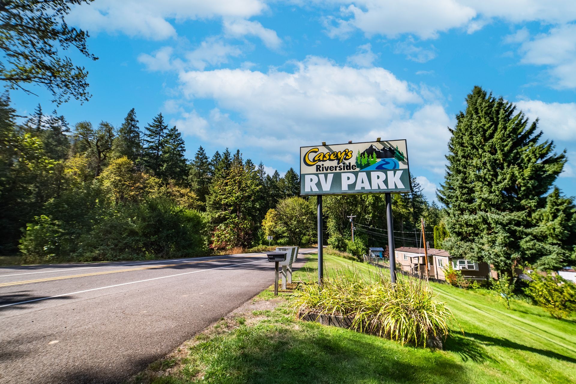 Road leading to an RV park sign, under a blue sky, trees in the background.