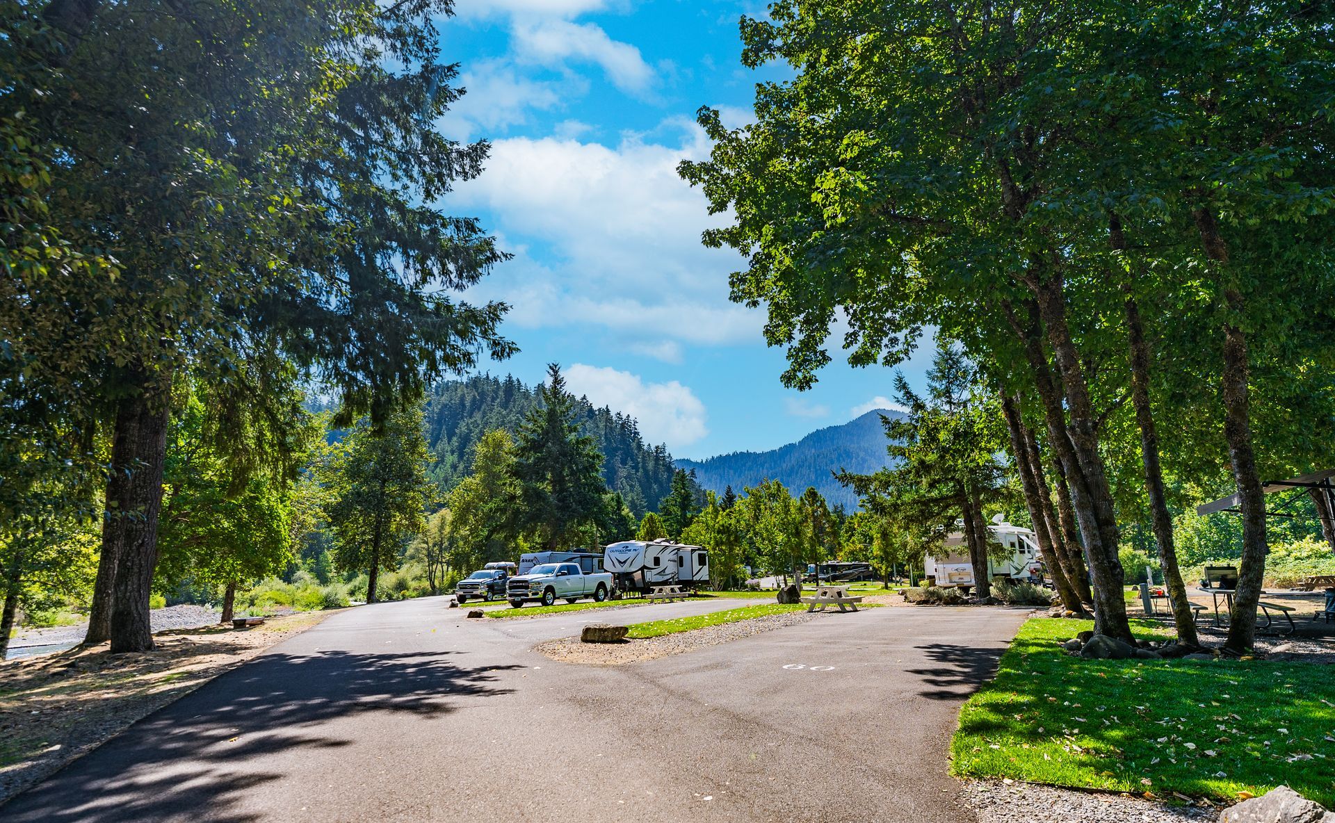 Campsite with RVs, trees, and mountains in the background under a partly cloudy sky.