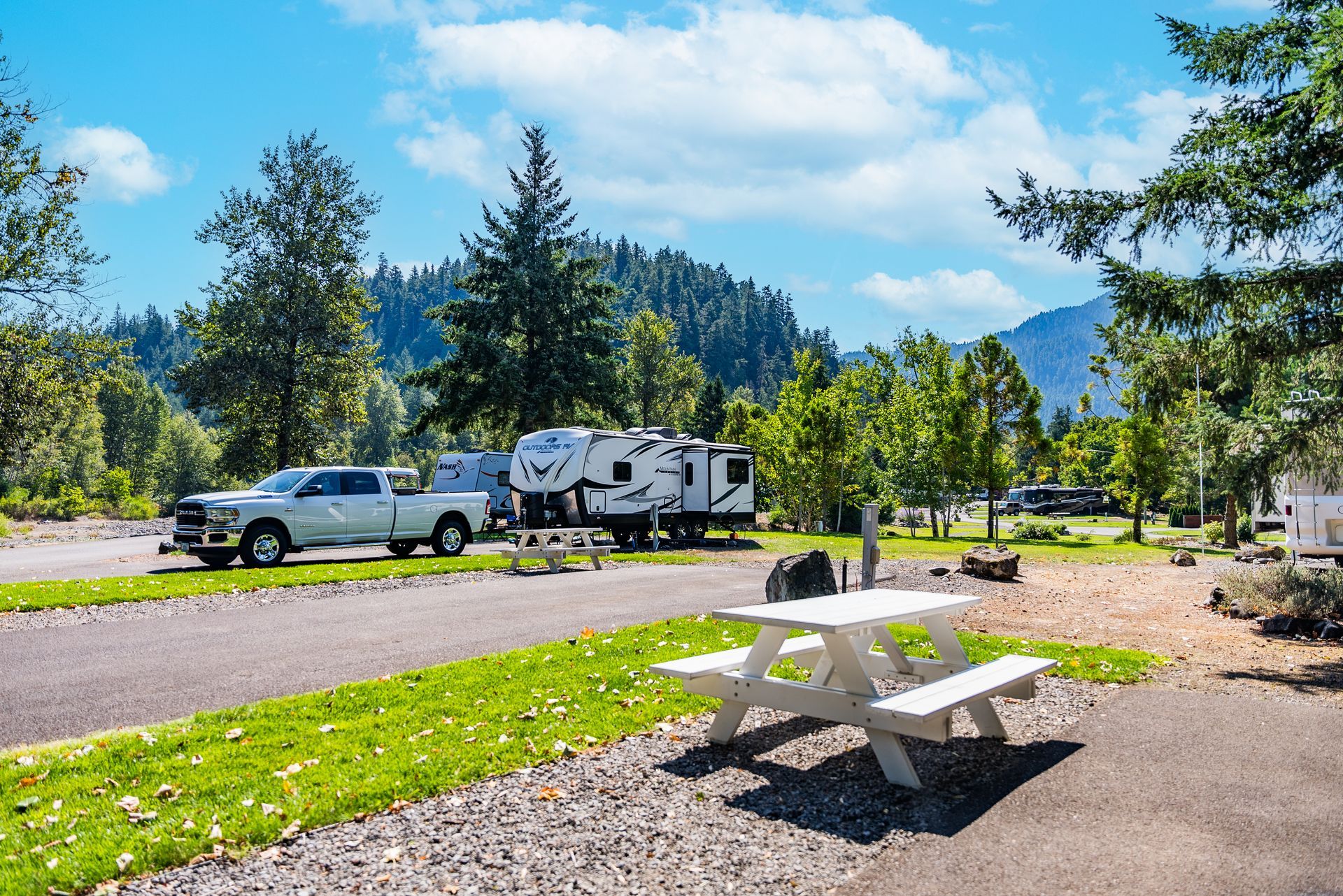 RV parked at a campground with a pickup truck, picnic table, and trees, mountains in the background.