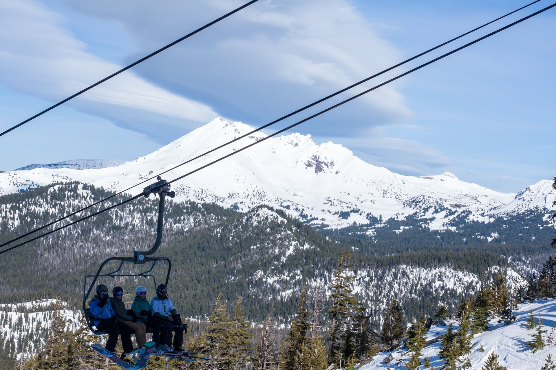 People ride a ski lift with a snowy mountain in the background. Sunny day with blue sky and some clouds.