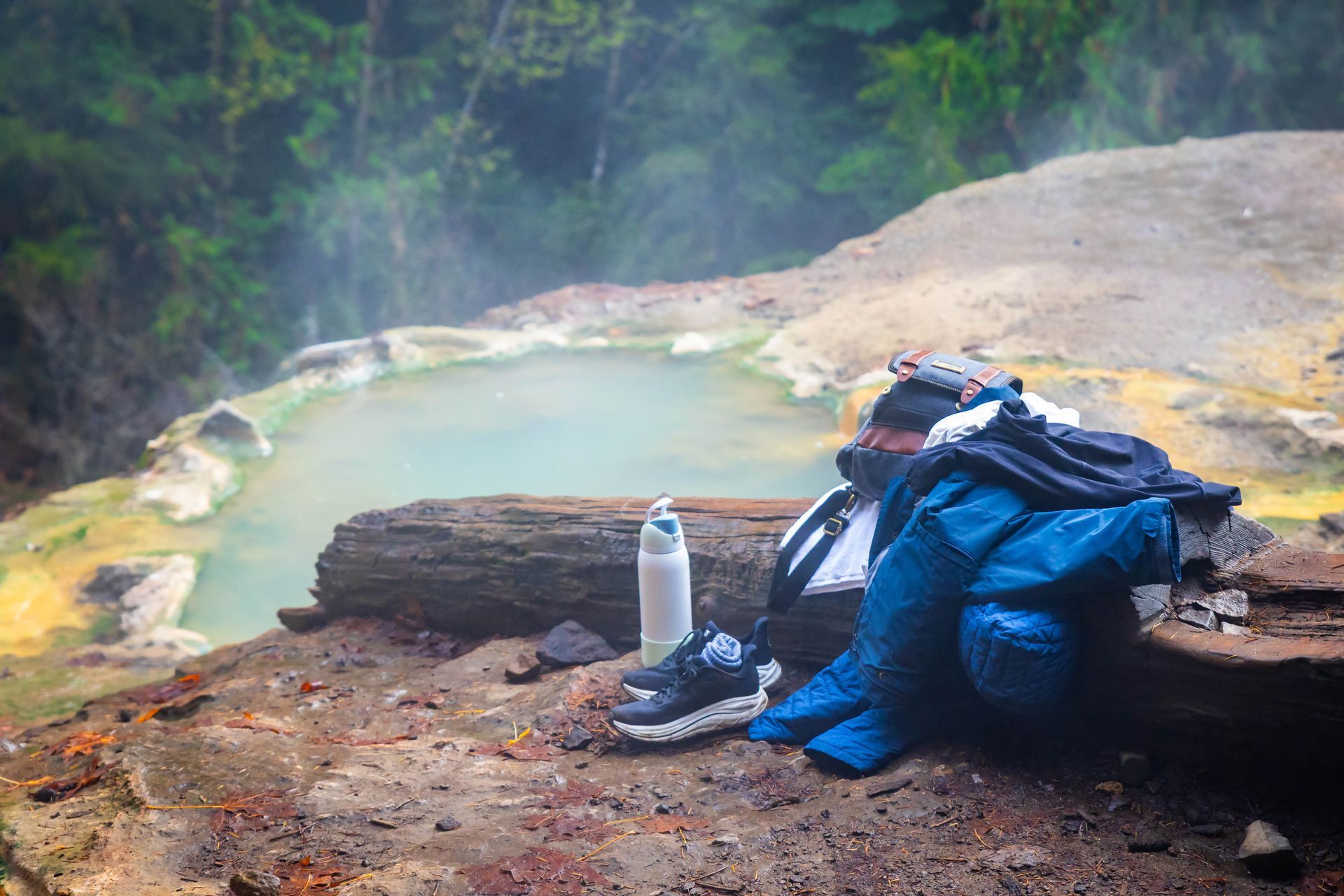 A person wearing a blue jacket rests against a log beside a steaming geothermal pool in a natural, rocky landscape.