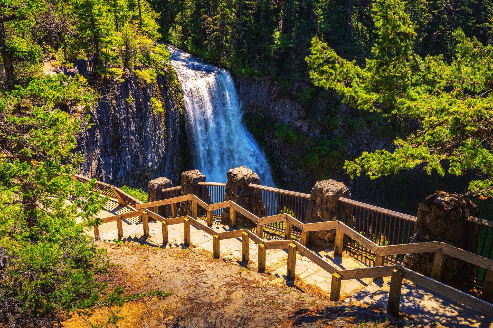 Waterfall cascading down a rocky cliff, viewed from a wooden staircase viewpoint, surrounded by trees.
