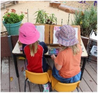 Two young children painting outdoors at a table, wearing hats.