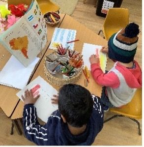 Two children drawing at a table with crayons, a book, and a basket of art supplies. One child wears a hat.