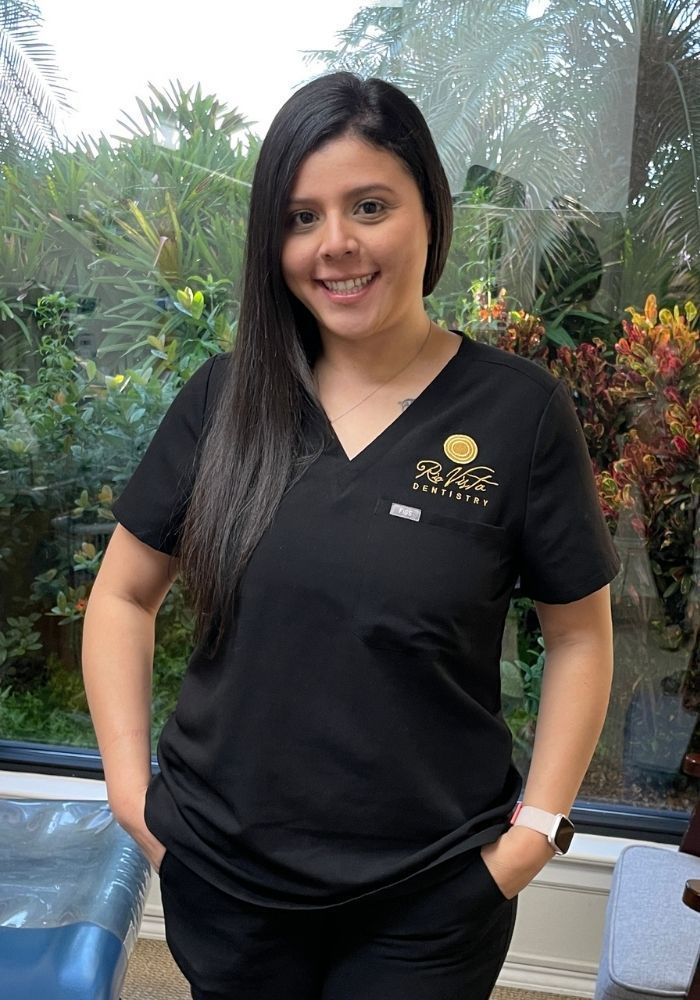 Woman in black scrubs, smiling, hands in pockets. Indoors, near a window with foliage.
