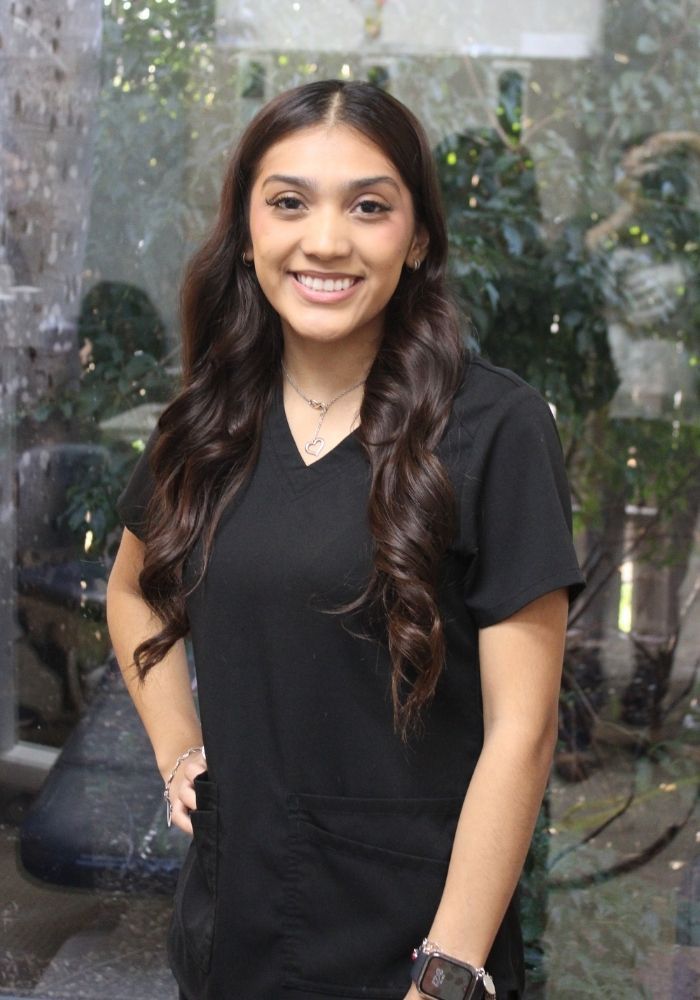 Woman in black scrubs smiling, standing in front of glass with greenery visible behind her.