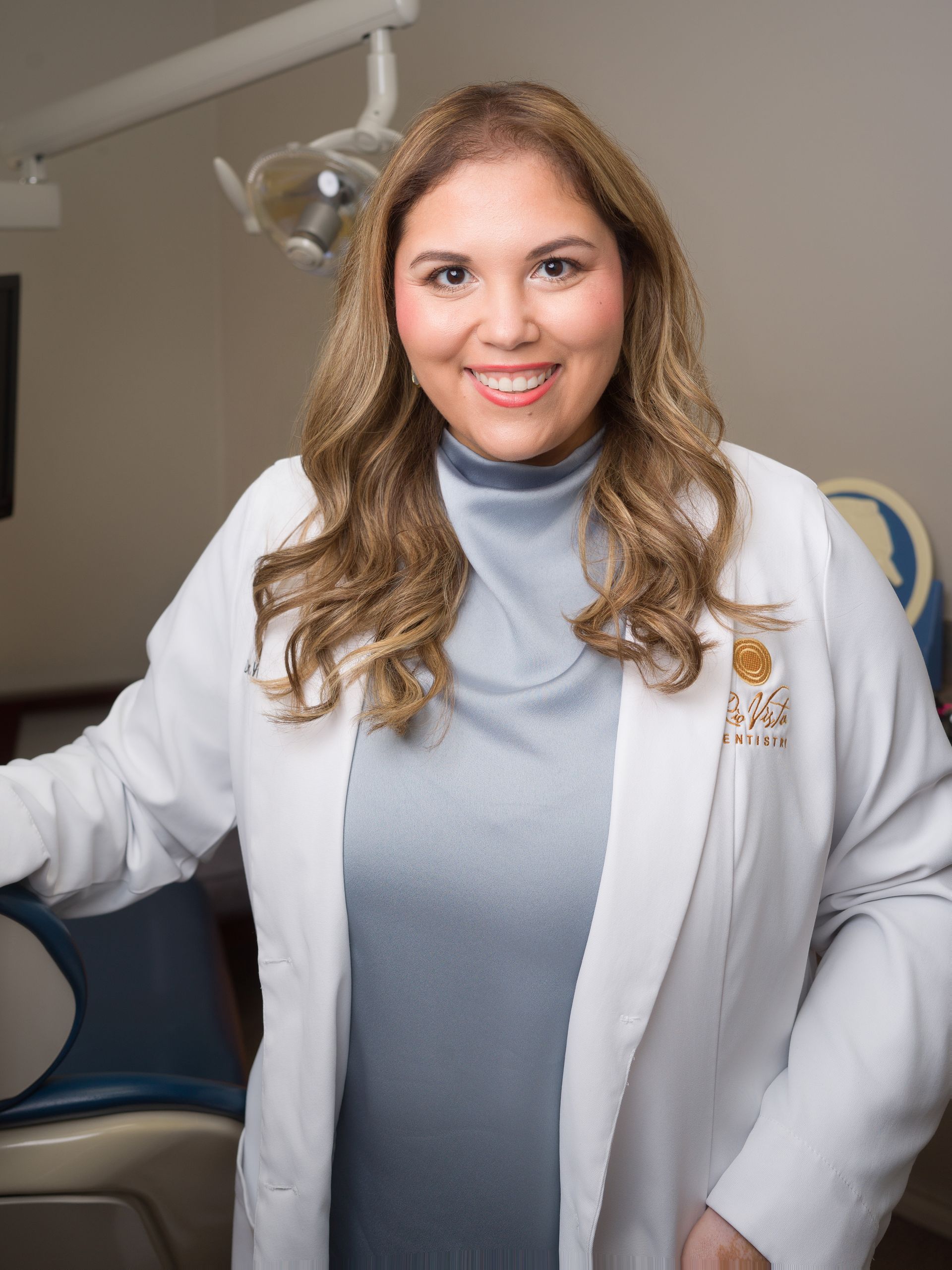 A professional in a white lab coat and blue top, smiling while standing in a dental office.