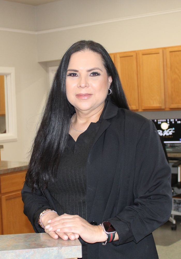 Woman with long dark hair, wearing a black blazer, standing in an office with wood cabinets.