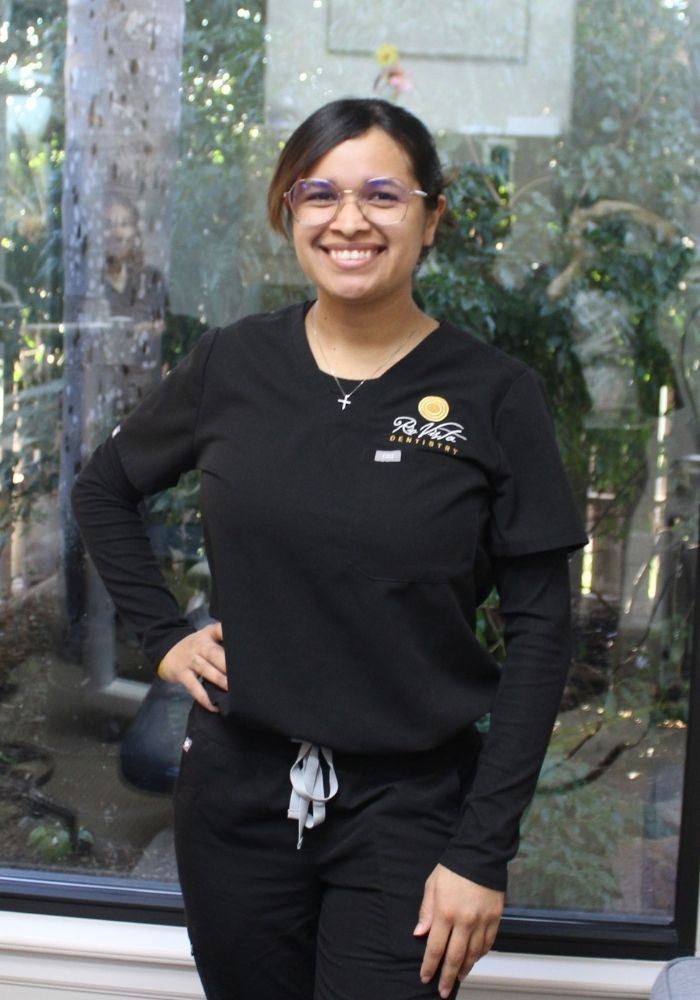 Woman in black scrubs, smiling, with hand on hip, near a window.