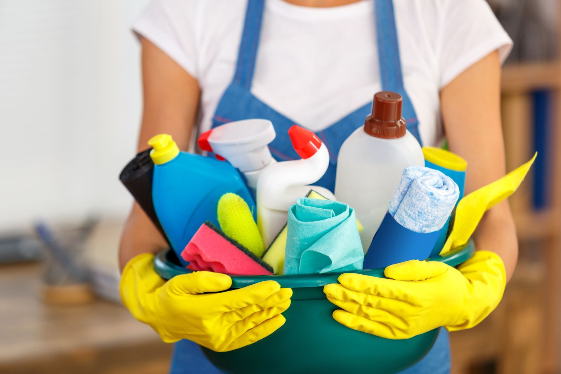 A person in yellow gloves holds a green basin filled with colorful cleaning bottles and cloths.