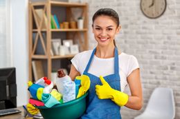 A smiling person in a blue apron and yellow gloves holds a basin of cleaning supplies, giving a thumbs up in an office. A smiling person in a blue apron and yellow gloves holds a basin of cleaning supplies, giving a thumbs up in an office.