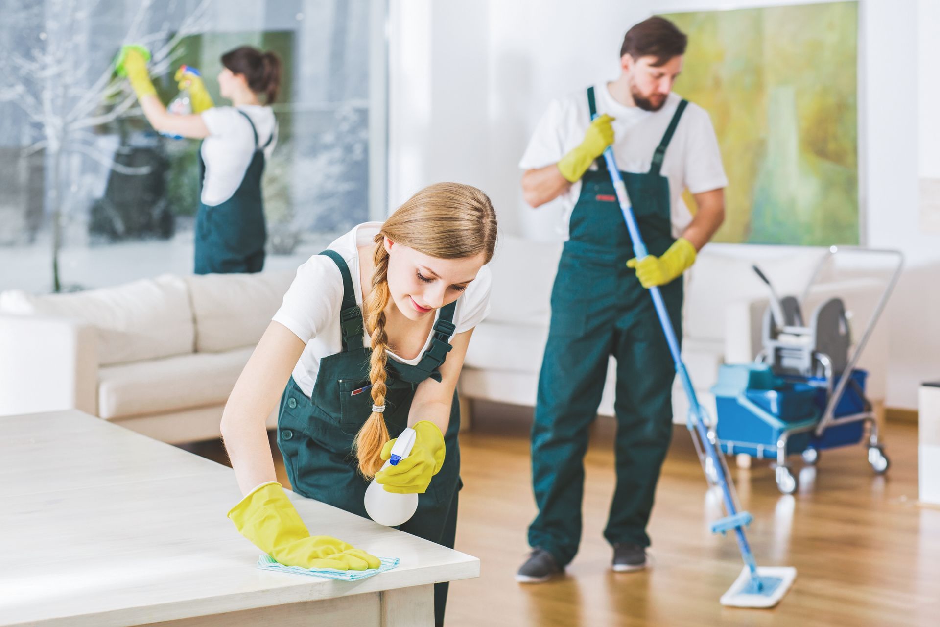 Three cleaning professionals in dark green uniforms work in a bright, modern living room. Three cleaning professionals in dark green uniforms work in a bright, modern living room.