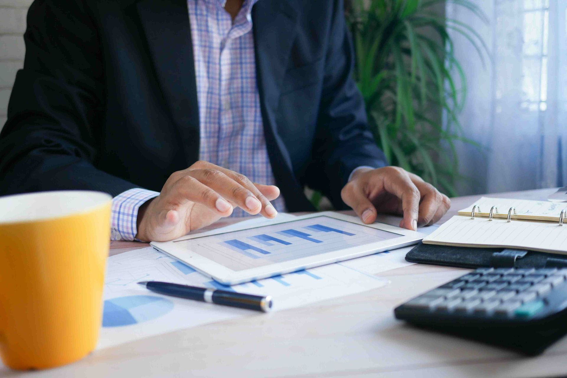 Person in blazer looking at tablet with charts, coffee mug, calculator, and notebook on desk.
