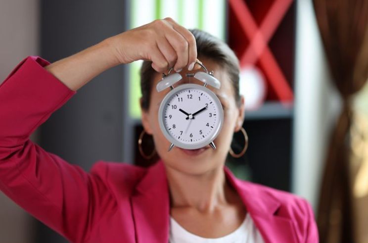 A woman in a pink jacket is holding an alarm clock in front of her face.
